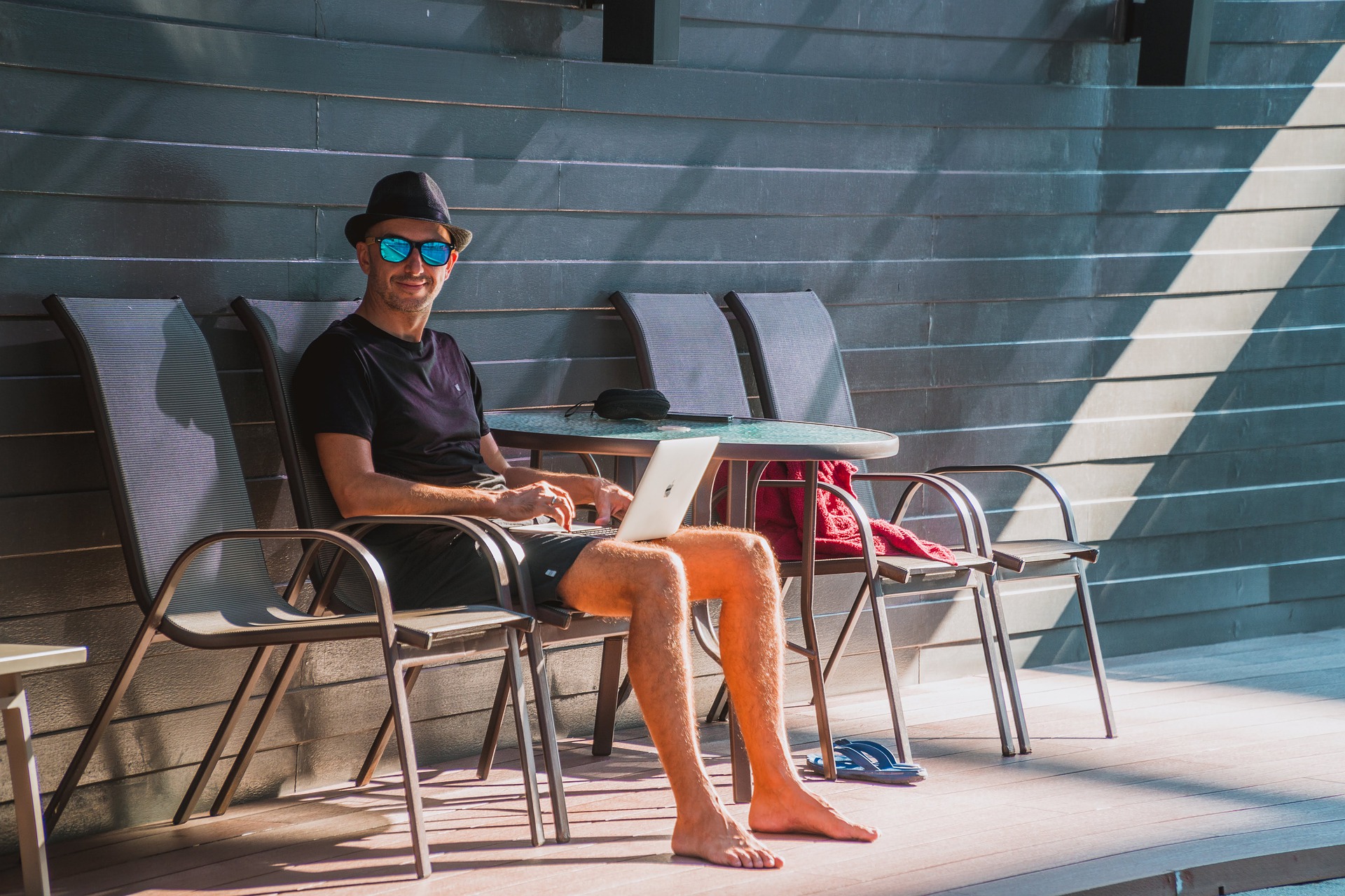 Homem descalço e com óculos de sol sentado em uma cadeira na beira da piscina trabalhando em seu notebook.