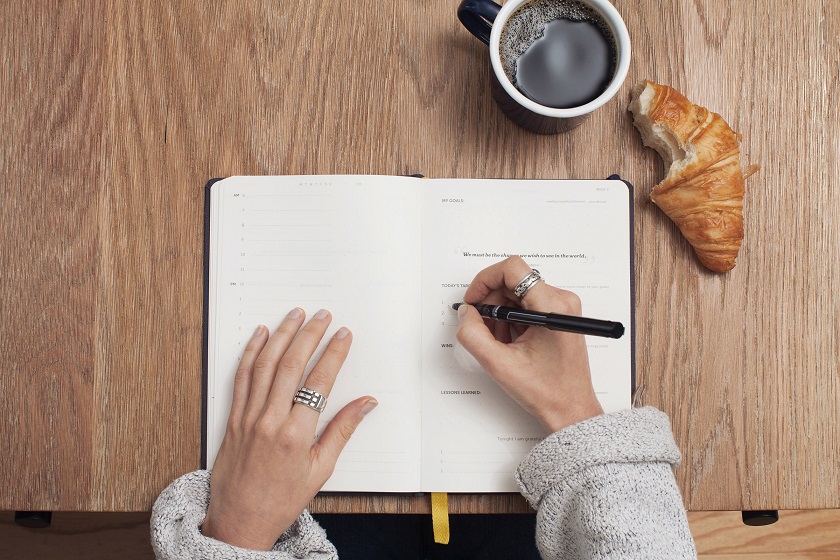 Foto de um caderno com uma mão realizando uma anotação, apoioado sobre uma mesa de madeira com uma xícara de café e um croissant.